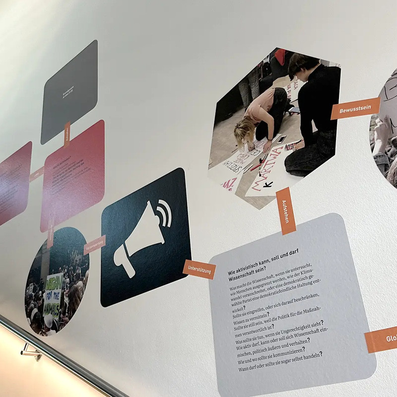 Wandinstallation entlang eines Treppengeländers. Geometrische Text- und Bildflächen sind wie eine Mindmap durch orange beschriftete Elemente miteinander verbunden. Zu sehen sind unter anderem Demonstrationsfotos und die Frage: "Wie aktivistisch kann, soll und darf Wissenschaft sein?"