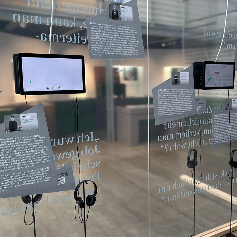Ausstellungsraum mit durchsichtigen beschrifteten Trennwänden. An den Wänden sind kleine Monitore mit jeweils darunterhängenden Kopfhörern angebracht.